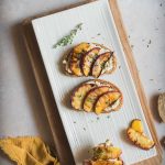 Crostoni di pane con ricotta di tofu e pesche grigliate su piatto rettangolare bianco e tagliere di legno