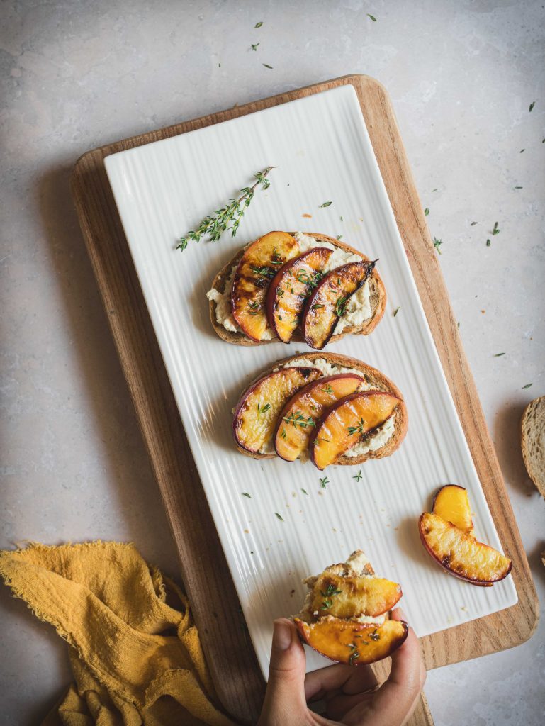 Crostoni di pane con ricotta di tofu e pesche grigliate su piatto rettangolare bianco e tagliere di legno